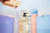 An assortment of Blueland cleaning products displayed against a light background. Includes toilet cleaner, hand soap, laundry detergent, and dishwasher tablets in pastel-colored containers. A hand is pouring powder from a brown pouch.