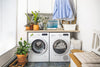Bright and organized laundry room with modern front-loading washer and dryer, potted houseplants, hanging shirts, and decorative storage on wooden countertop
