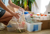 Person sorting recyclables into reusable containers, including glass jars, with a mesh shopping bag on the floor.