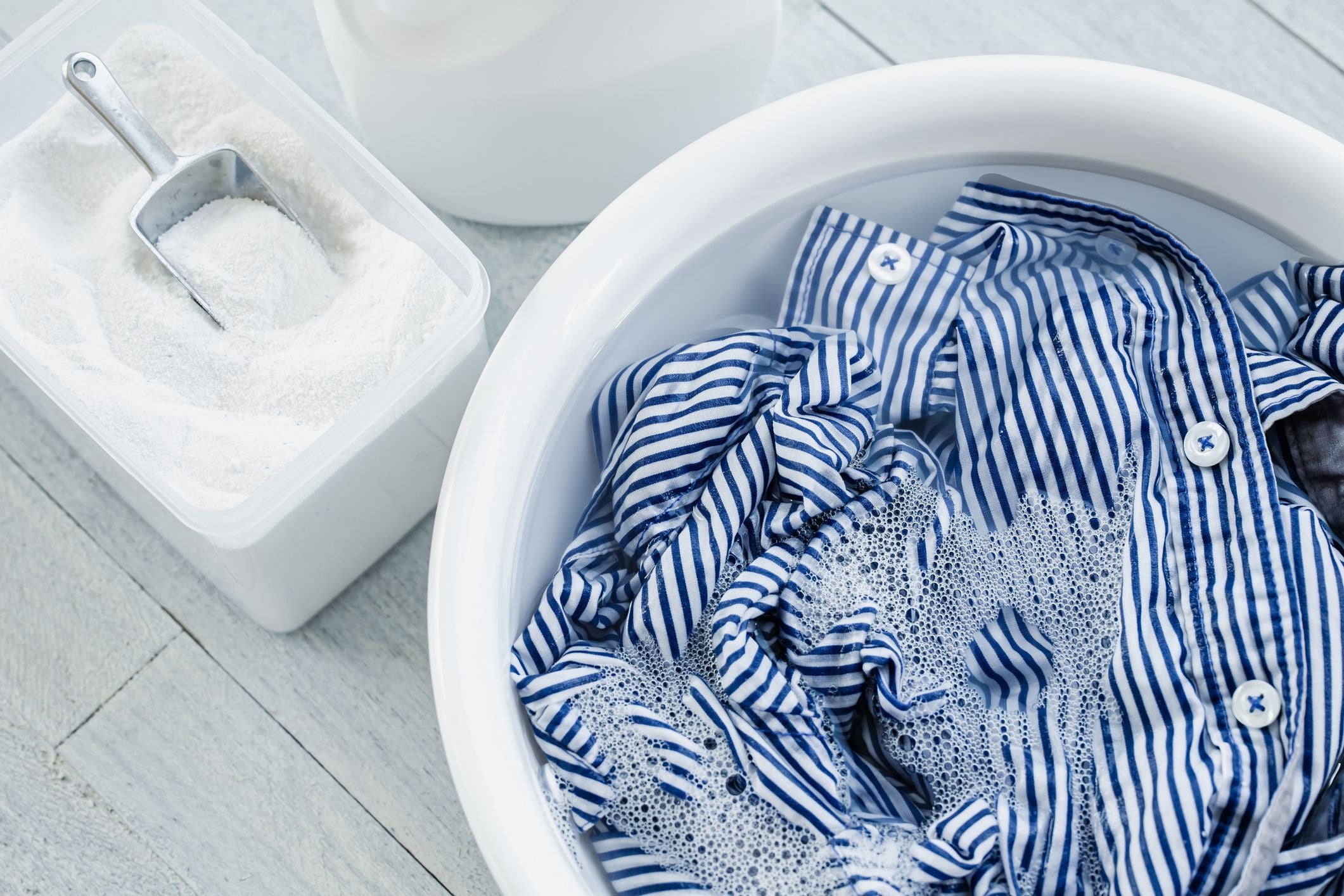 Blue and white striped shirt soaking in a white bucket with soap, on a light wooden floor.