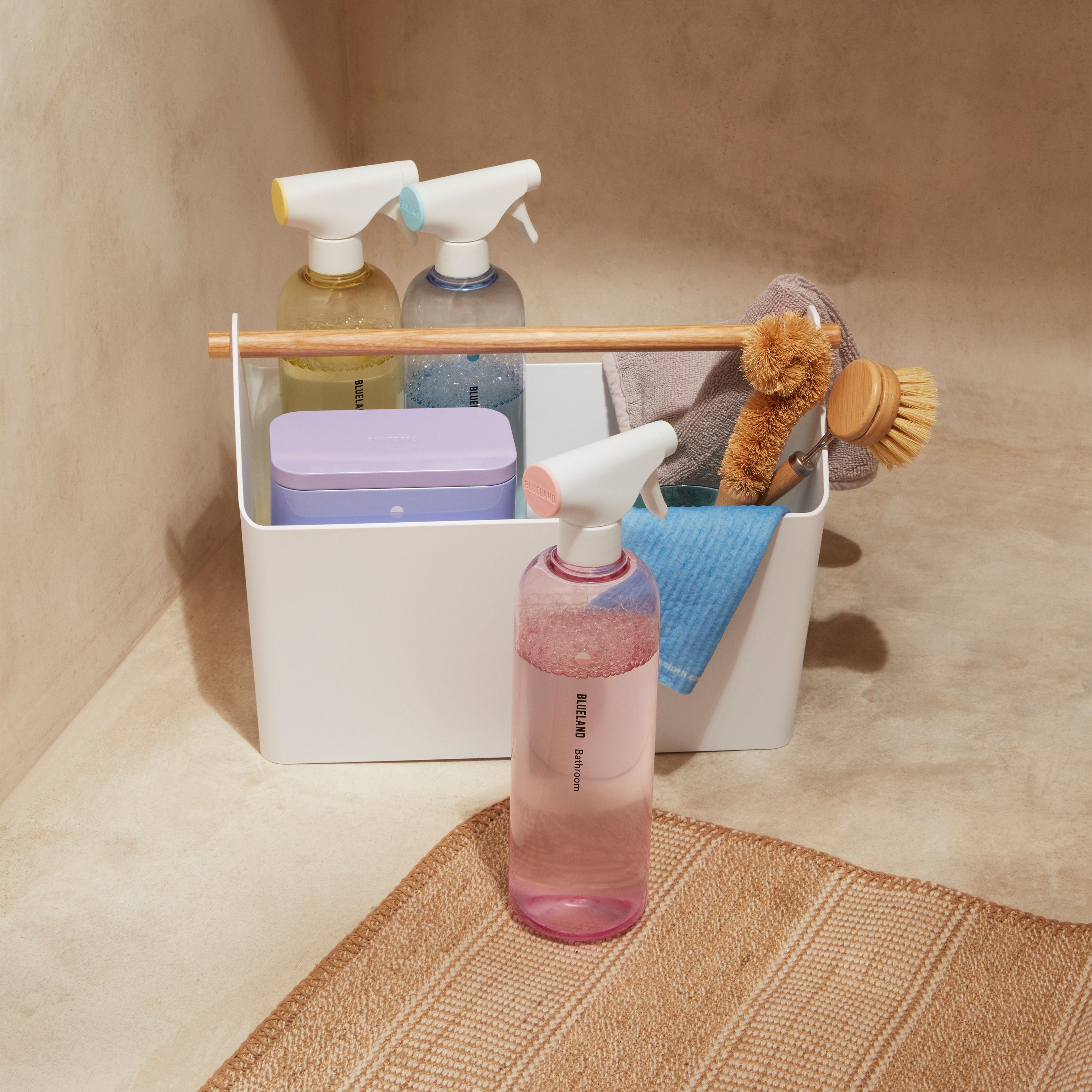A white storage caddy holds spray bottles, a lavender box, a scrubbing brush, a sponge, and a blue cloth. A pink spray bottle stands outside the caddy on a woven mat in a beige room.