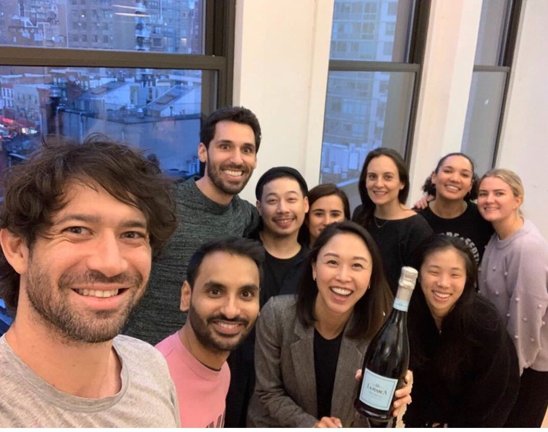 A group of ten people smile for a selfie indoors near large windows, with a woman in front holding a bottle of sparkling wine. City buildings are visible outside the windows.